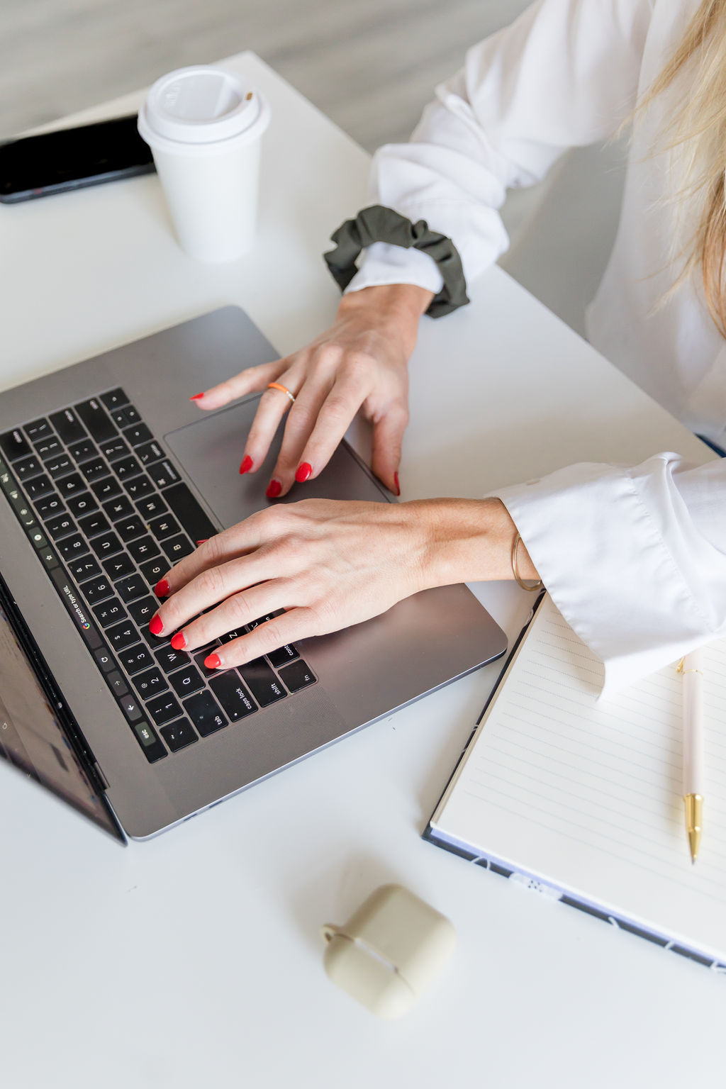 Woman in white shirt typing on a laptop on a white desk next to coffee headphones and a notebook